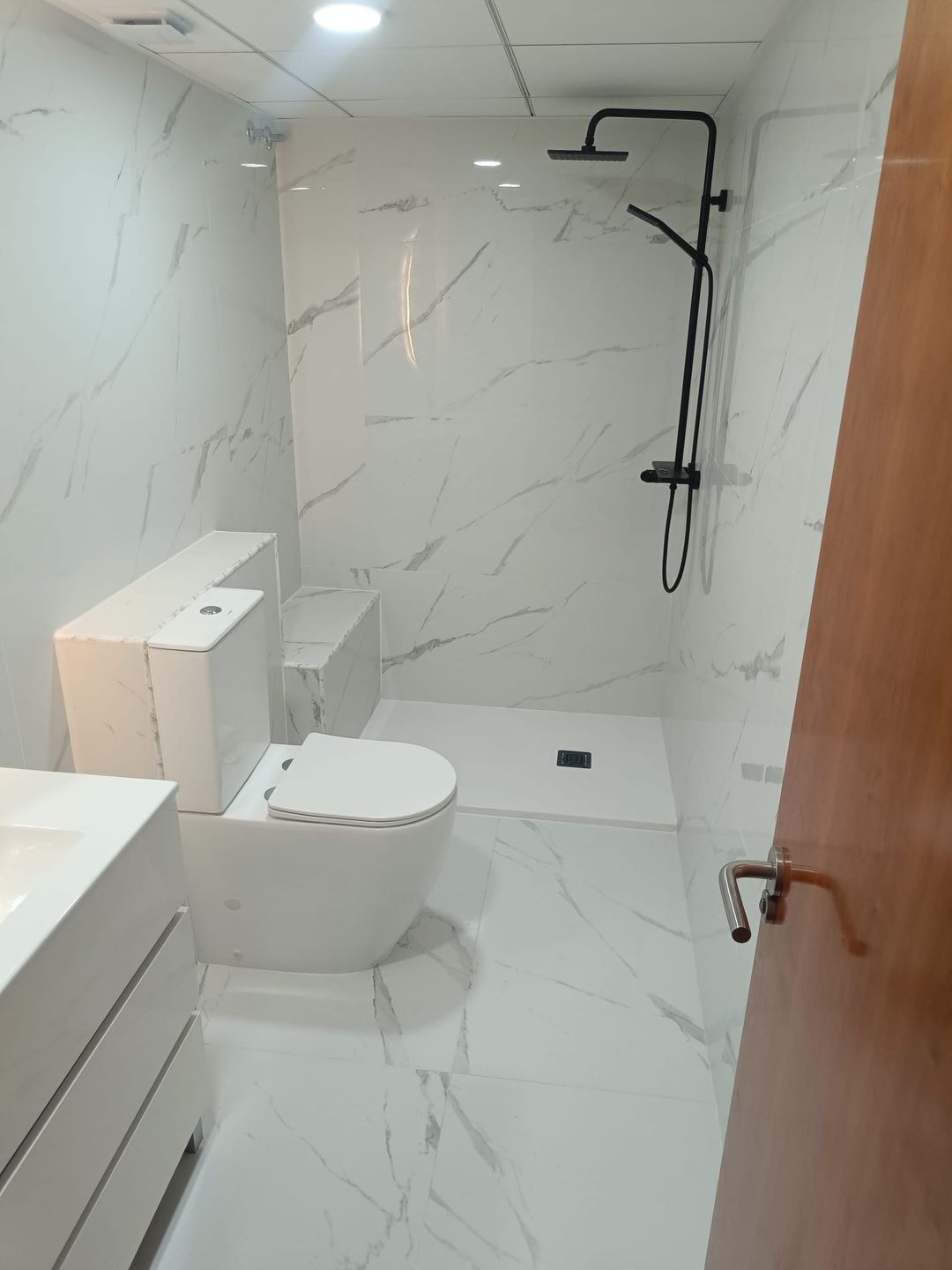 Baño blanco de mármol con plato de ducha a ras de suelo, columna negra e inodoro
