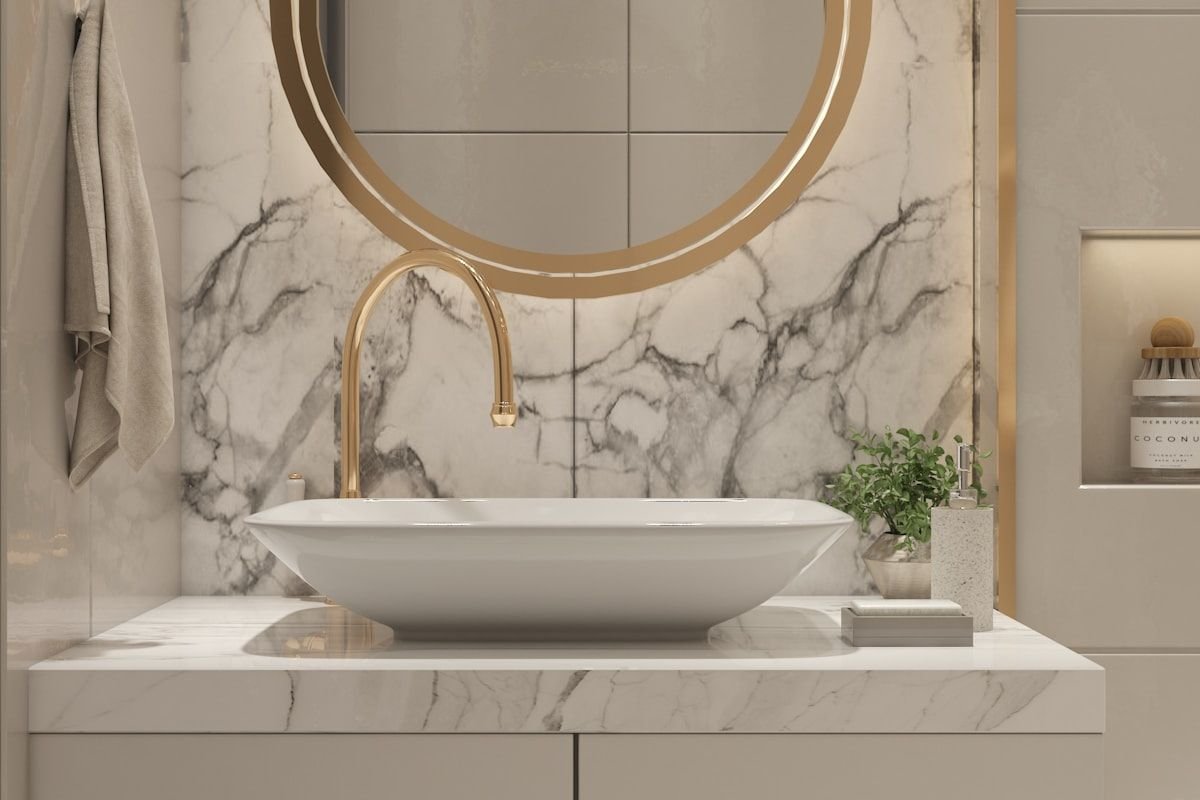 Baño de mármol claro con lavabo sobre encimera y espejo redondo con marco dorado