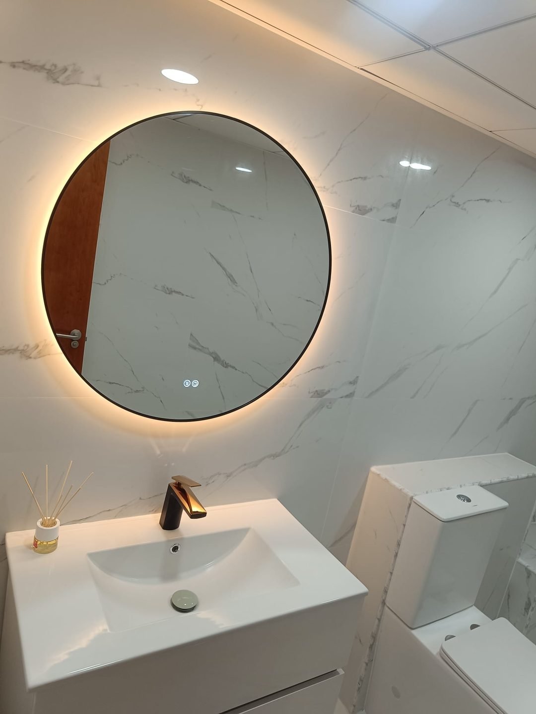Mueble de baño blanco con grifería en negro y oro rosa y espejo redondo retroiluminado de aro negro