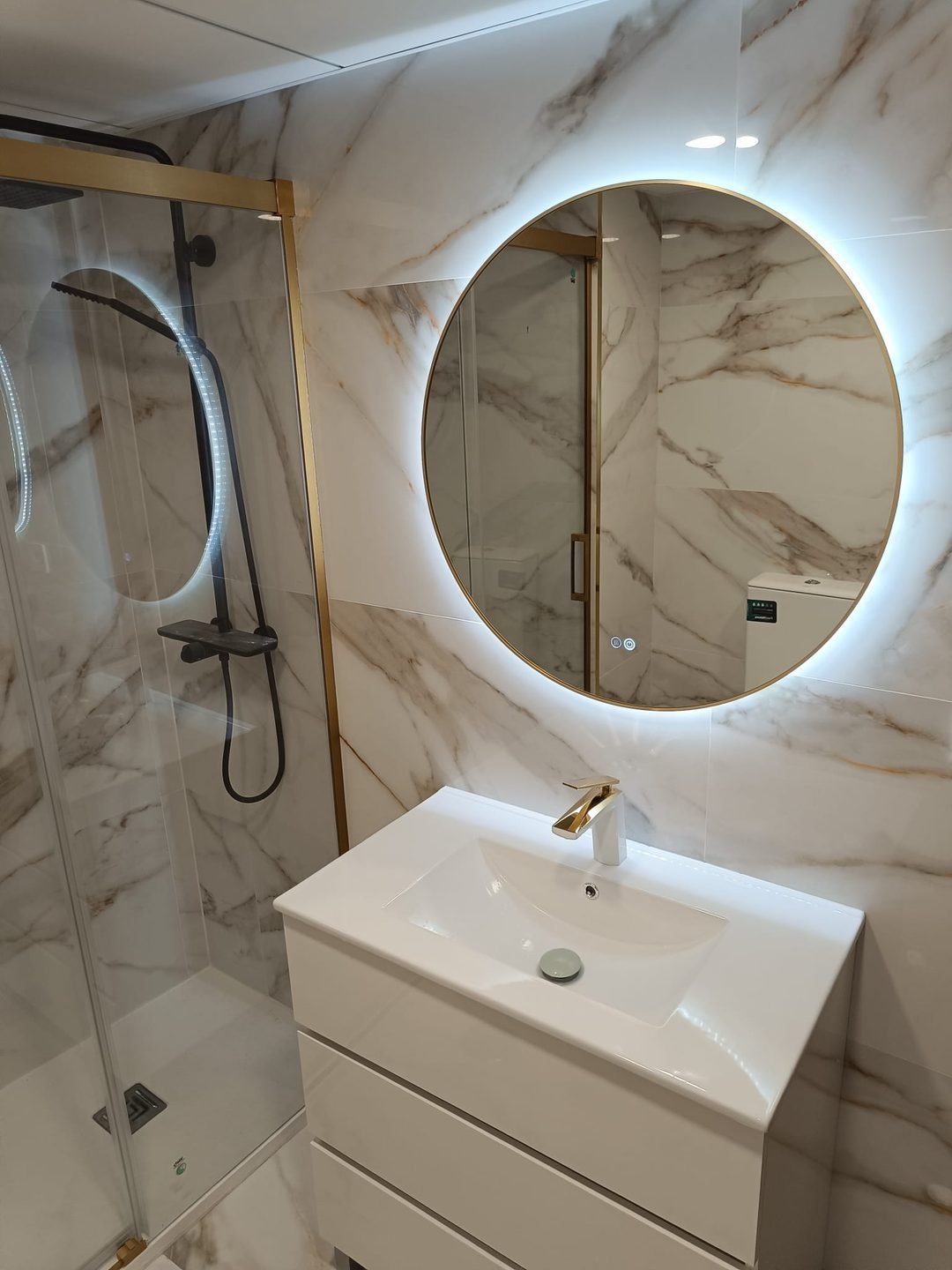 Baño terminado con mampara corredera de perfil dorado, espejo redondo retroiluminado y grifería dorada