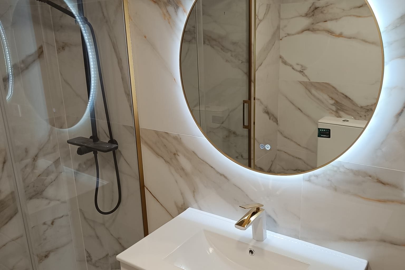 Baño reformado con mampara de perfil dorado y espejo redondo retroiluminado