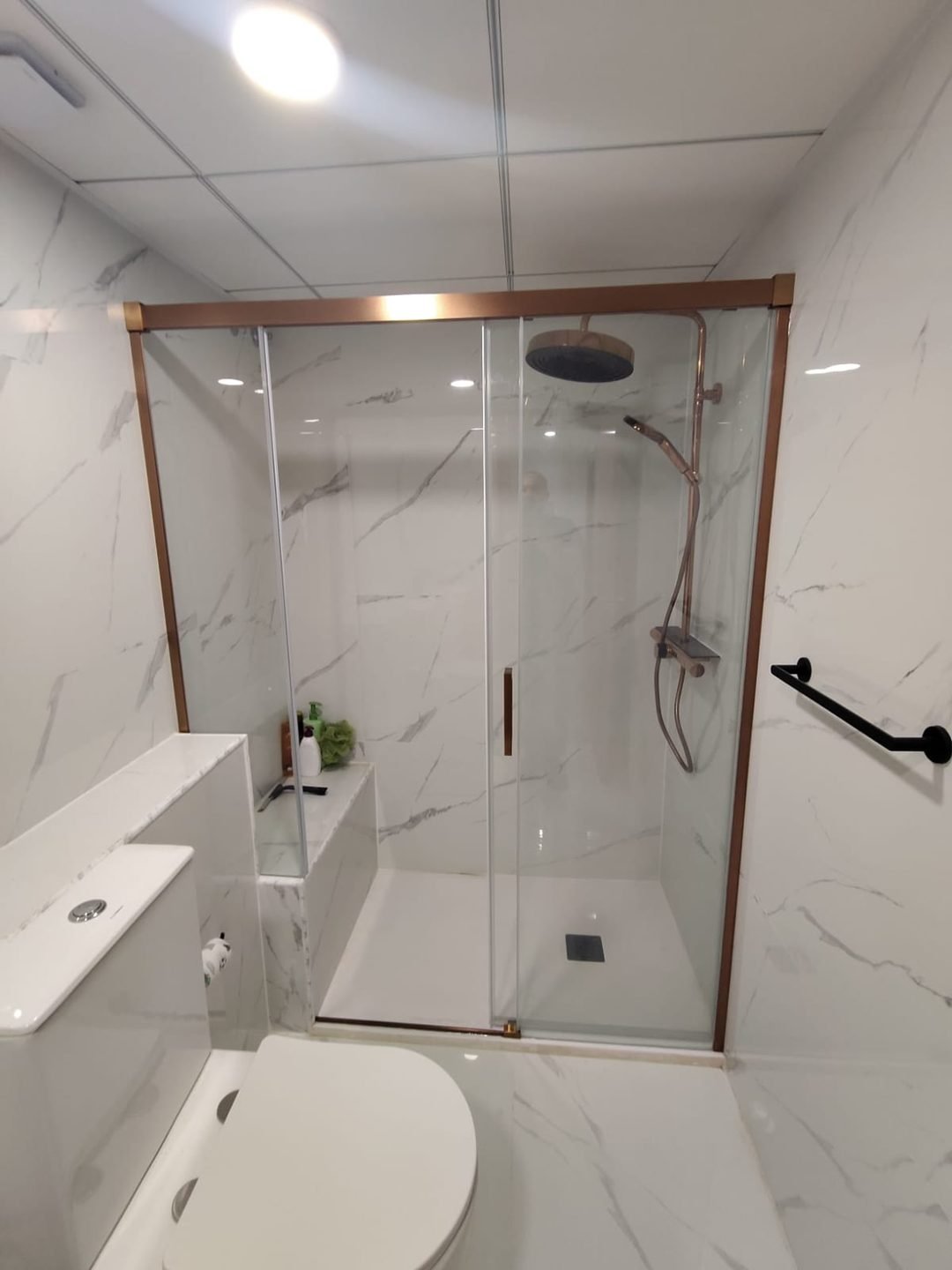 Ducha terminada con mampara de tres paños y perfil en oro rosa, banco de obra alicatado y columna a juego