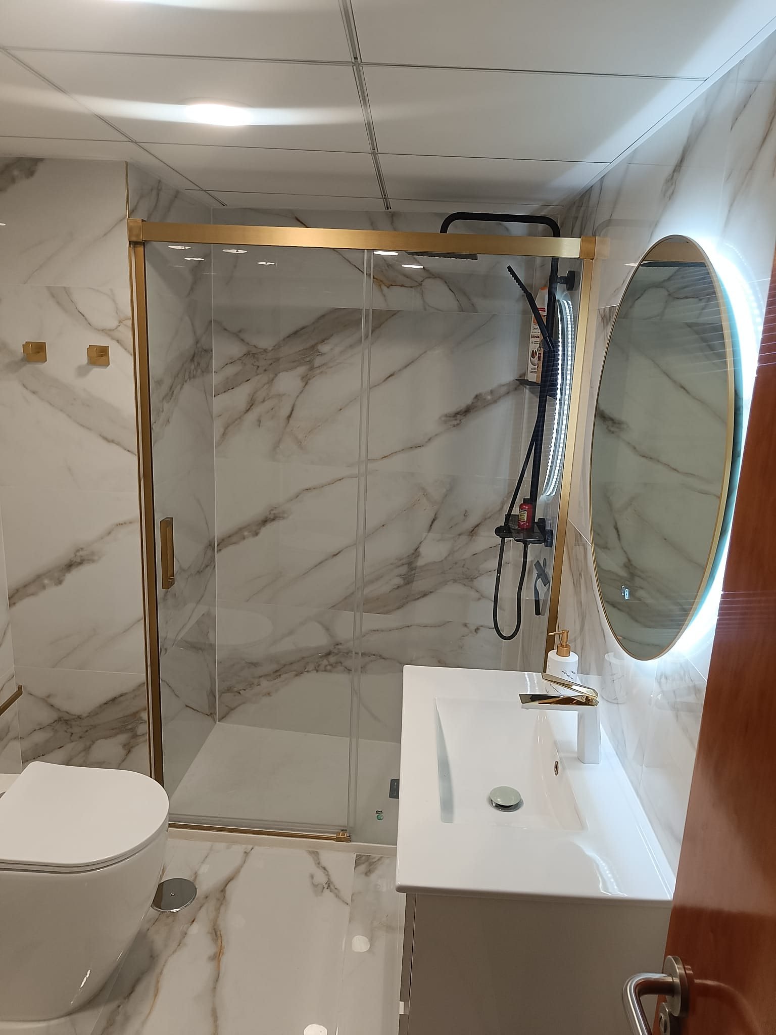 Baño entregado en Alicante: porcelánico efecto mármol, mampara de perfil dorado, columna de ducha negra, espejo redondo retroiluminado y grifería dorada