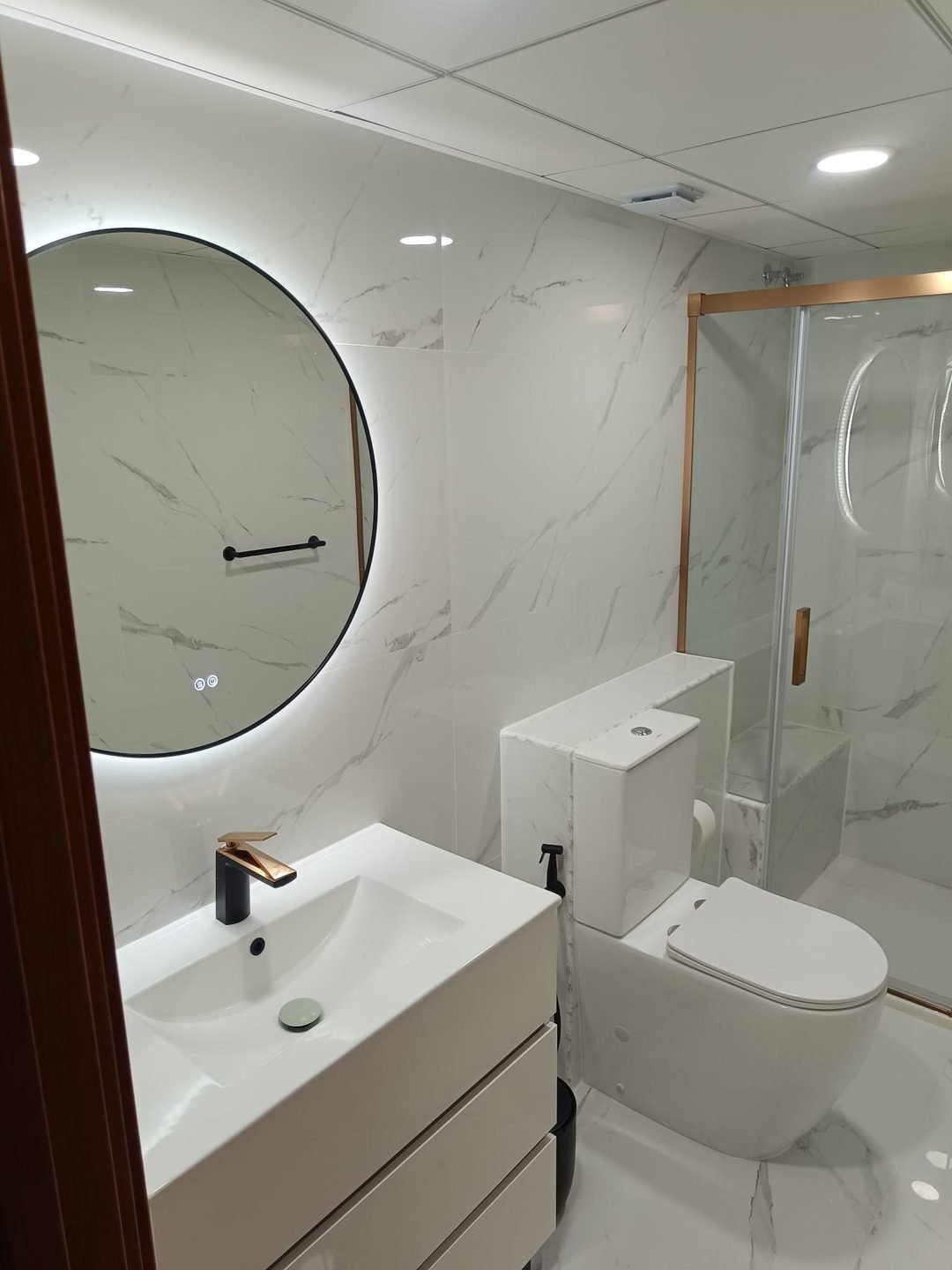 General view of the finished bathroom, with the round backlit mirror, vanity unit, WC and the shower at the back