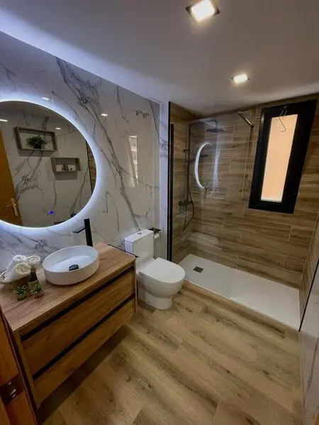Baño moderno con espejo redondo iluminado, lavabo de cerámica y ducha de cristal.